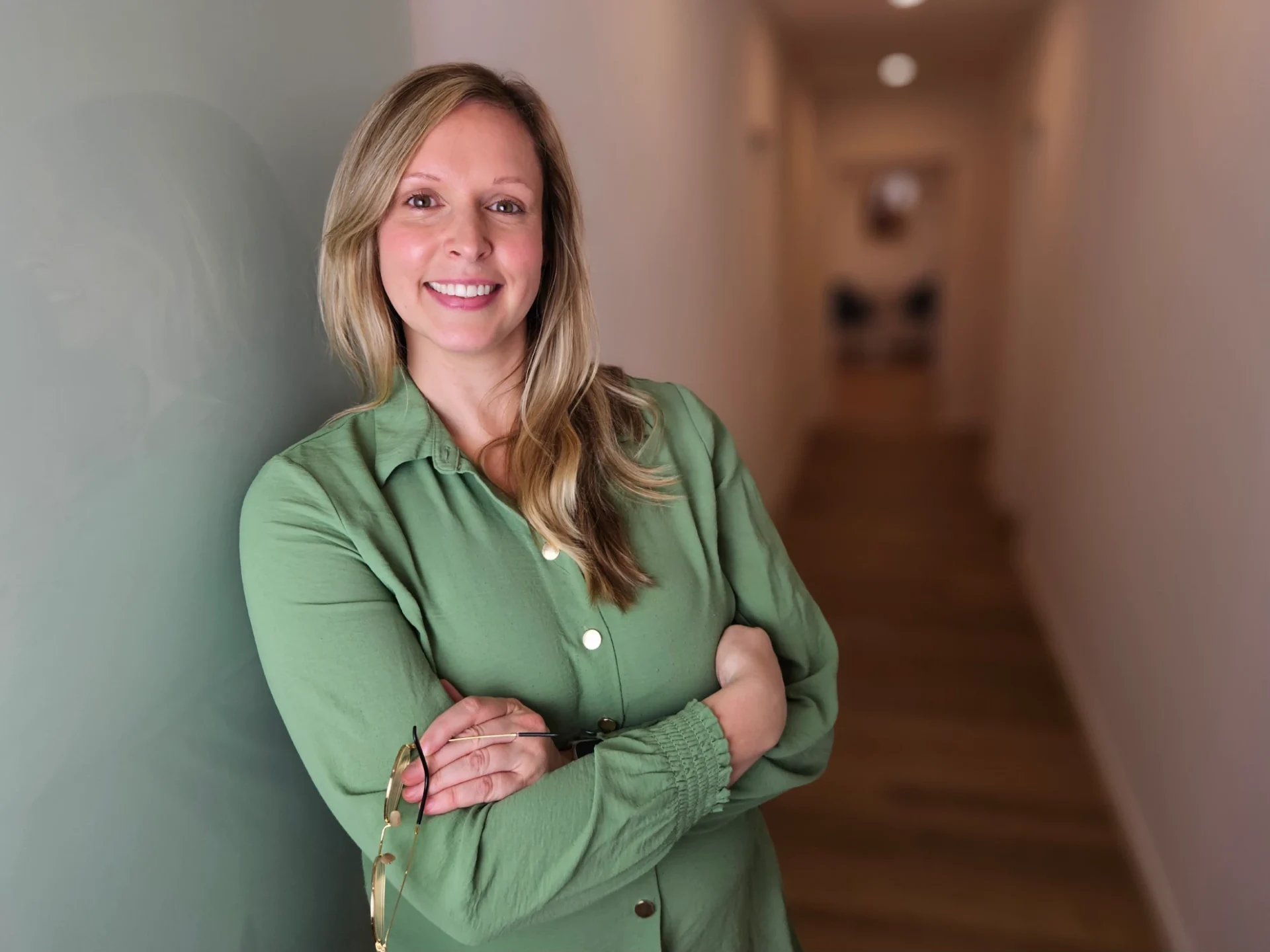 Porträt einer sympathischen Frau mit blonden Haaren, grünem Hemd, lehnt an einer grünen Wand in einem Flur; freundlicher Gesichtsausdruck, Mentacura-Umgebung im Hintergrund