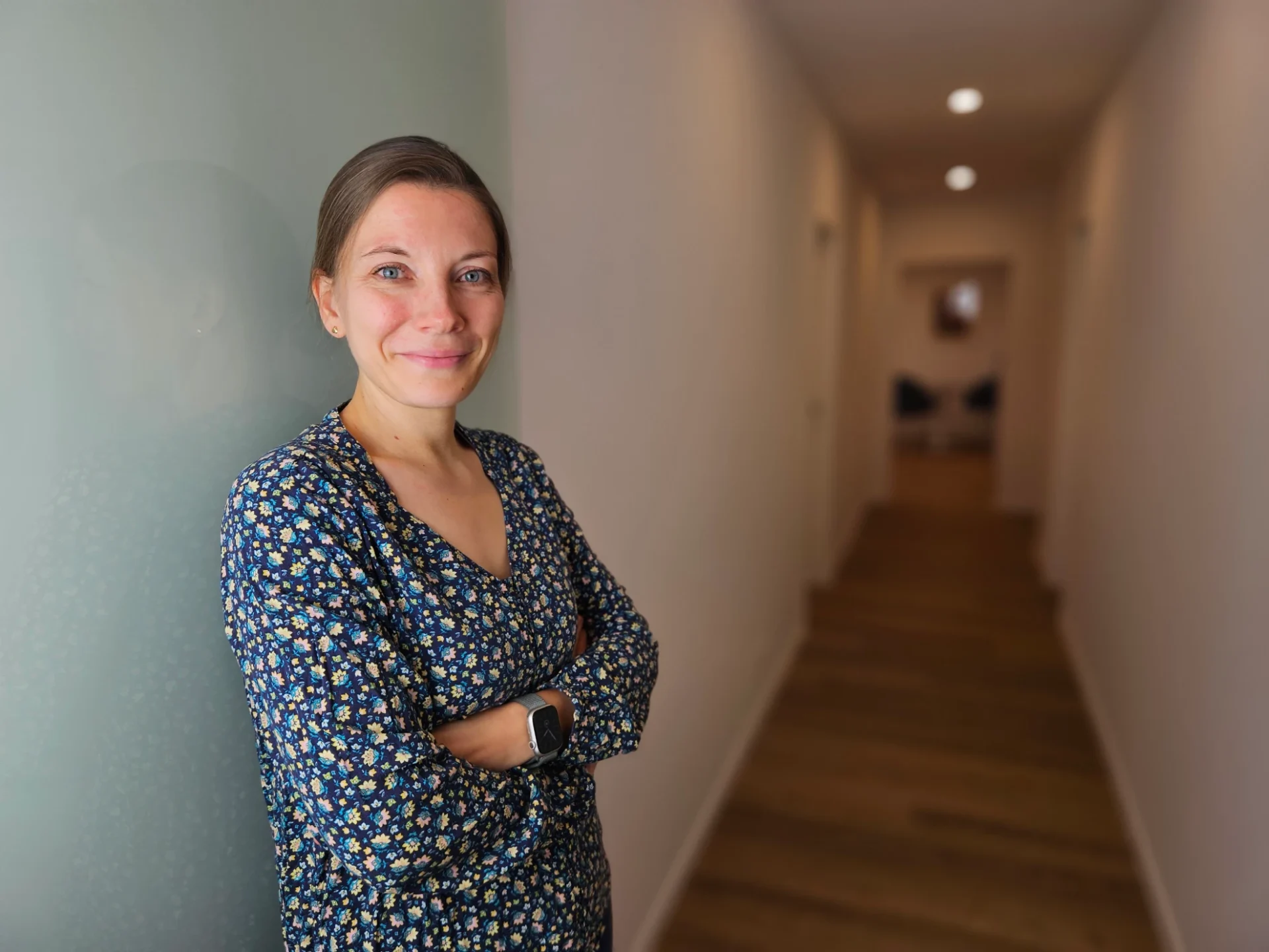 Portrait einer Frau mit dunkelblauem Blumenmuster-Kleid, lächelnd, die Arme verschränkt an einer grünlich gestrichenen Wand in einem hellen Flur stehend.