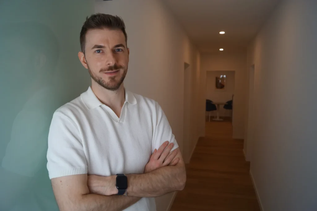 Portrait eines Mannes in einem hellen Flur, trägt ein weißes kurzärmliges Polohemd und eine schwarze Armbanduhr; Blick in die Kamera, Mentacura Umgebung im Hintergrund.